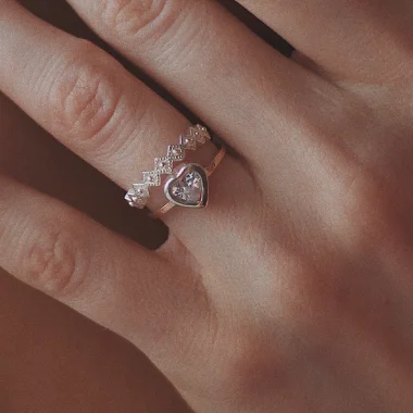 Detalle de dos anillos finos en la mano: uno de líneas en zigzag con brillos y otro con piedra en forma de corazón.