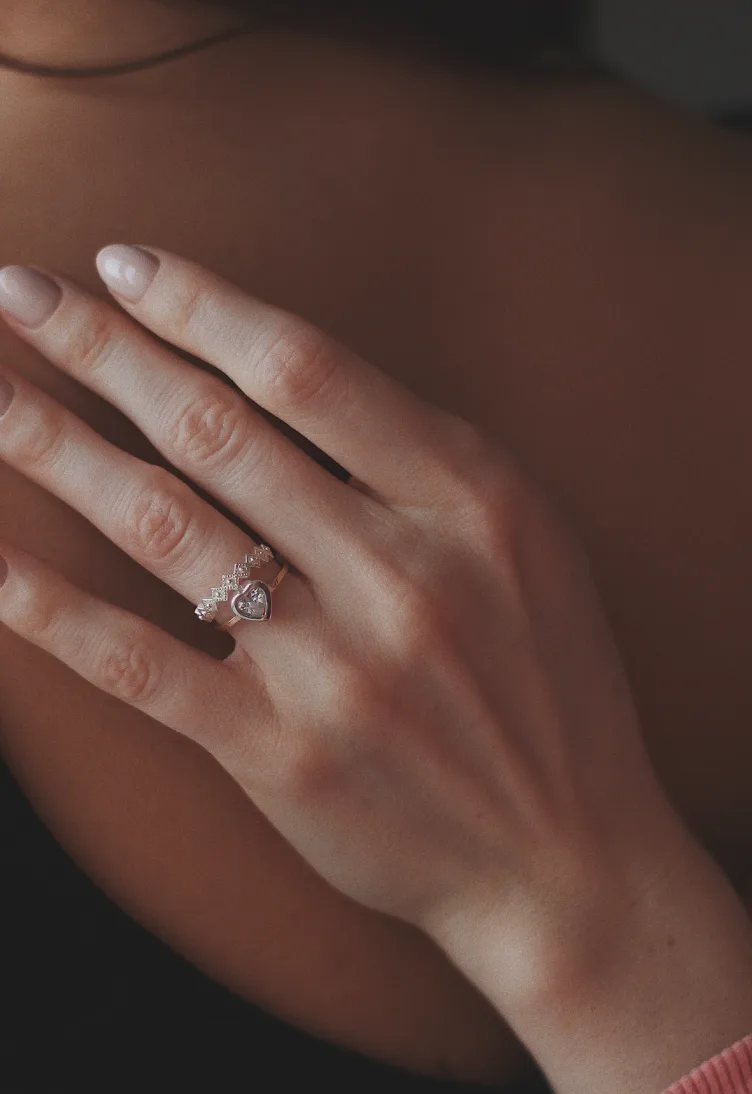 Mano apoyada sobre el hombro con dos anillos finos, uno con piedra en forma de corazón, en luz cálida y tenue.